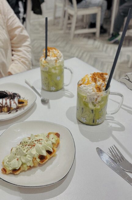 Zwei Gläser Matcha mit Sahne und knusprige Croffles im Café Always Together – ein farbenfroher Genussmoment im schwarz-weißen Interior.