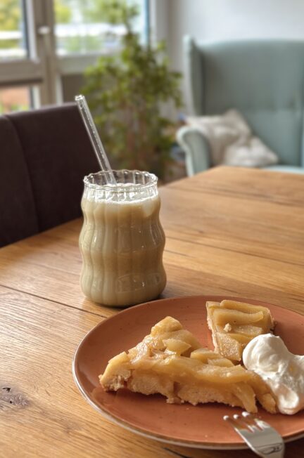 Ein Glas Matcha-Latte mit Glasstrohhalm neben einem Teller mit Birnen-Tarte und Sahne auf einem Holztisch im gemütlichen Wohnzimmer.