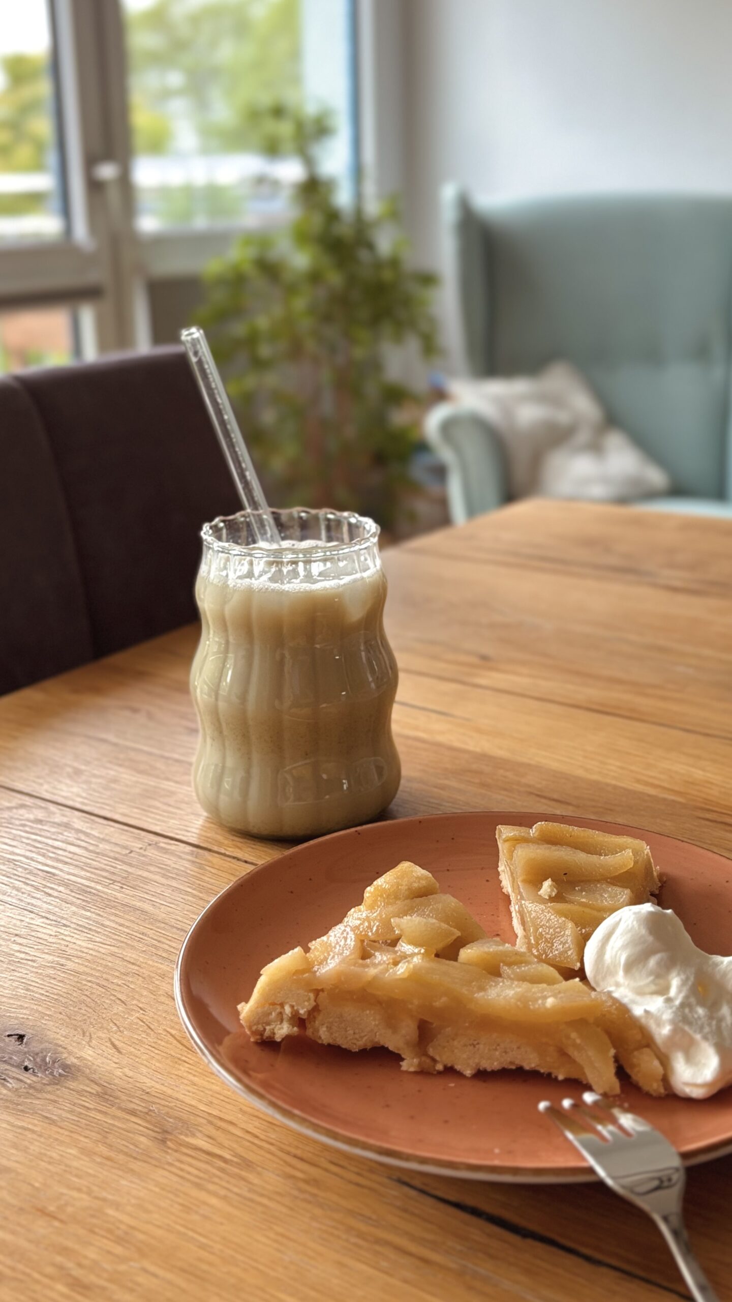 Ein Glas Matcha-Latte mit Glasstrohhalm neben einem Teller mit Birnen-Tarte und Sahne auf einem Holztisch im gemütlichen Wohnzimmer.